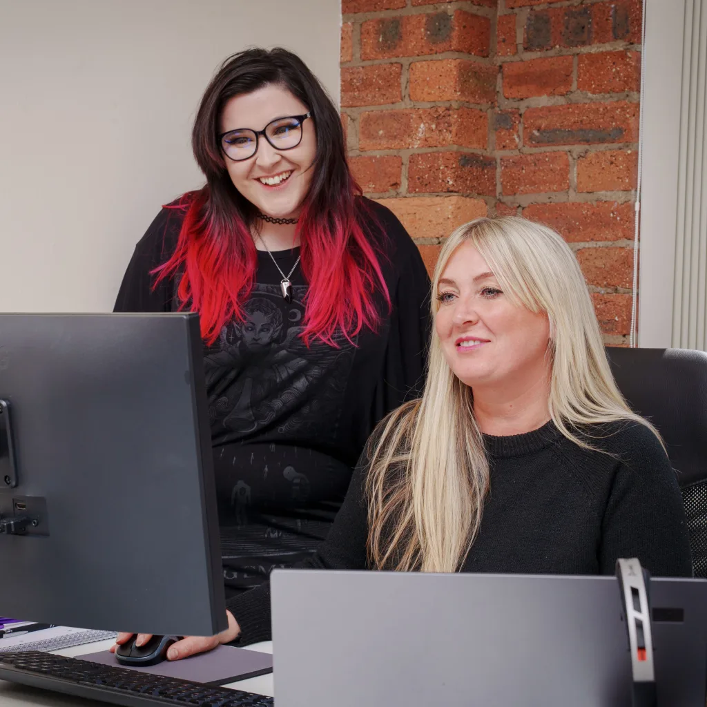 Two women at a desk with computers, collaborating and discussing their work in a professional setting.