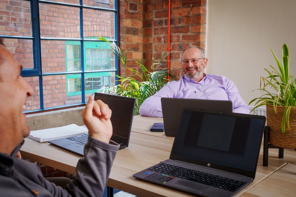 Two smiling men sitting at a table with laptops during a meeting.