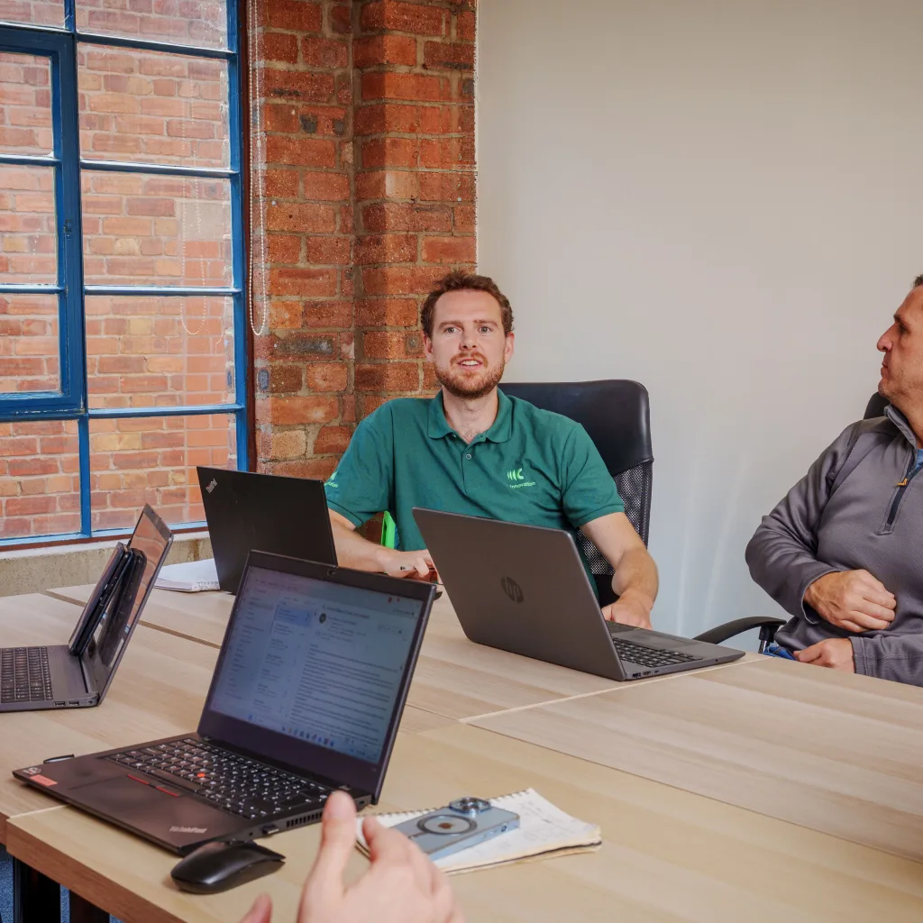 A man wearing a Cyd Innovation uniform is seated at a table with three laptops, engaged in a meeting.