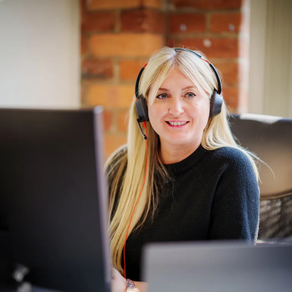 A woman wearing a headset sits at her desk, smiling as she answers phone calls.