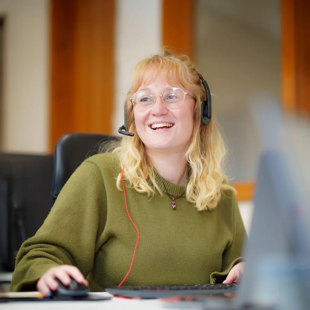 A smiling woman in a green sweater answers phones while working at her computer.