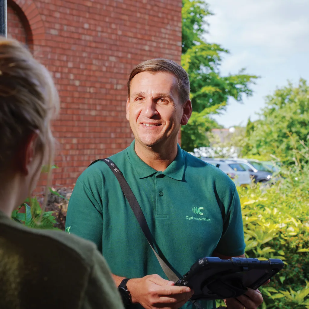 A man in a green shirt stands at the door, greeting a woman and holding a digital device.